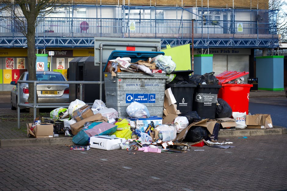 An outdoor scene showing multiple overflowing rubbish bins and scattered waste on a paved area near a parking space. The primary object is a large, metal recycling bin marked for mixed paper and cardb