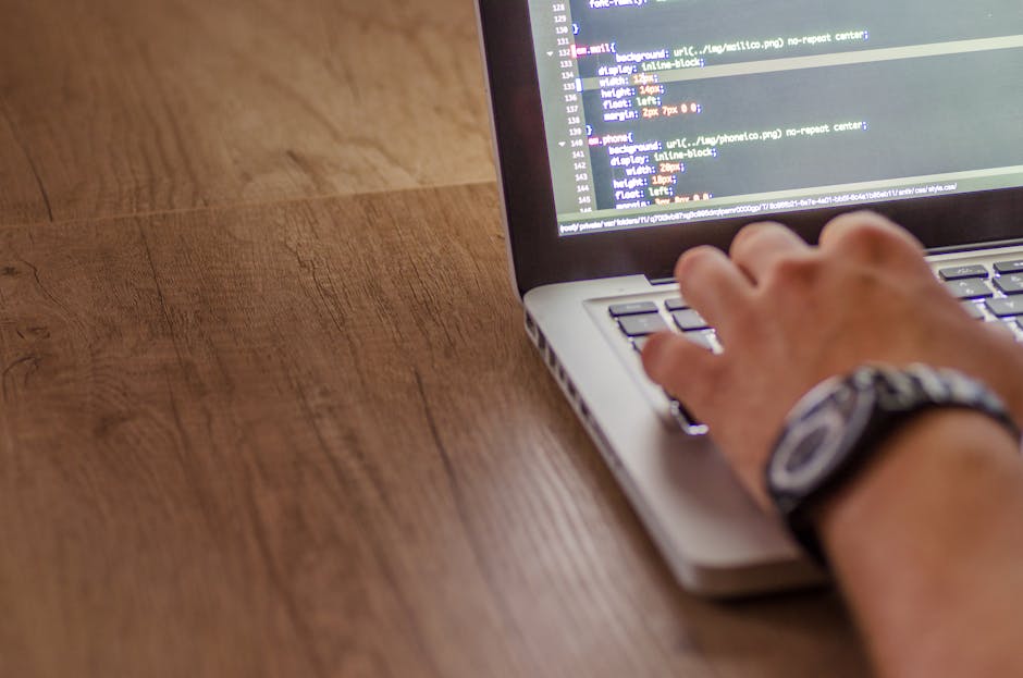 A close-up view of a person's hand using a silver laptop with a black keyboard and a screen displaying lines of code or programming text. The hand, wearing a wristwatch with a dark band, is positioned