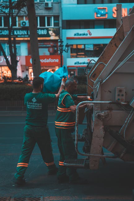 A worker dressed in green uniform and wearing a safety helmet stands on the rear platform of a small, hooded rubbish collection truck, which is parked on a suburban street with a slight incline. The t